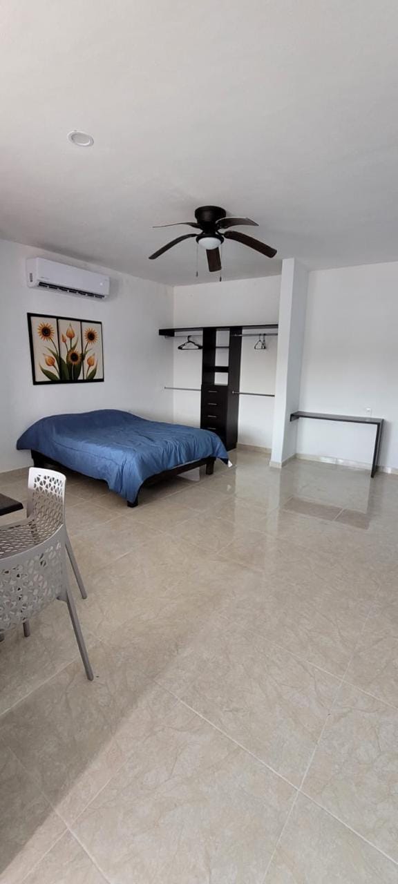 A spacious sleeping area features a blue bedspread on a large bed. A ceiling fan provides airflow. A modern dresser and hangers for clothing are visible, along with a small dining table accompanied by two chairs. A floral artwork adds a touch of color to the wall.
