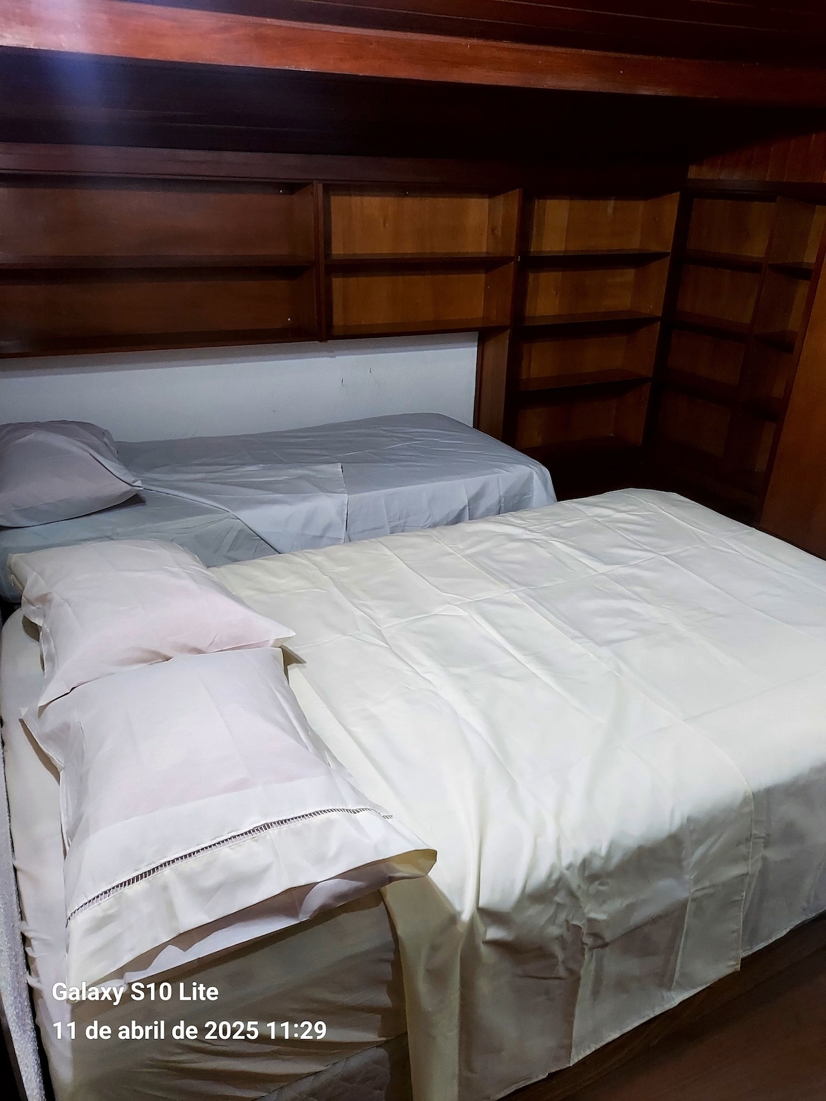 A cozy sleeping area features two beds, one with a light-colored blanket and the other with a smooth, white coverlet. Wooden shelves line the walls, providing ample space for personal items. Soft light enhances the warm tones of the wooden furnishings.