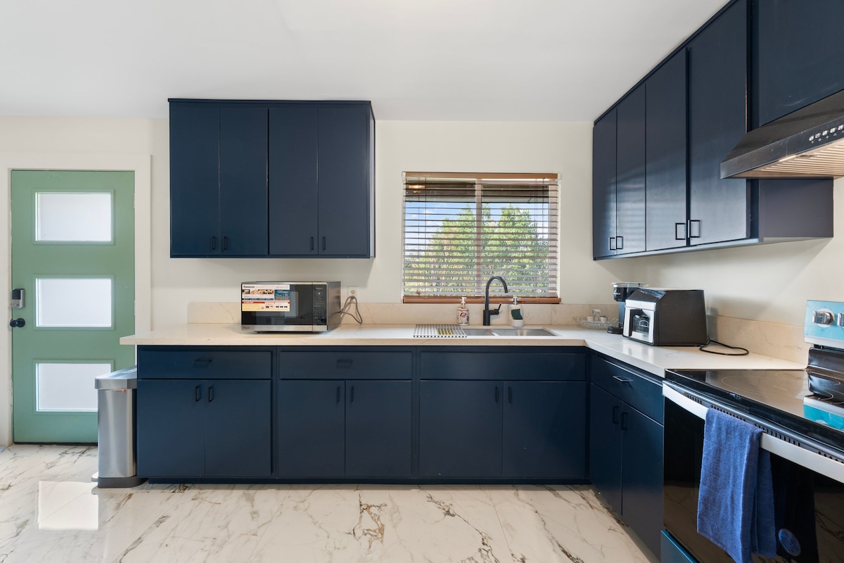 A modern kitchen features dark blue cabinetry and light marble countertops. An organized layout includes a sink, microwave, and stovetop. A window allows natural light to enter, while a door with a glass panel leads to the exterior.