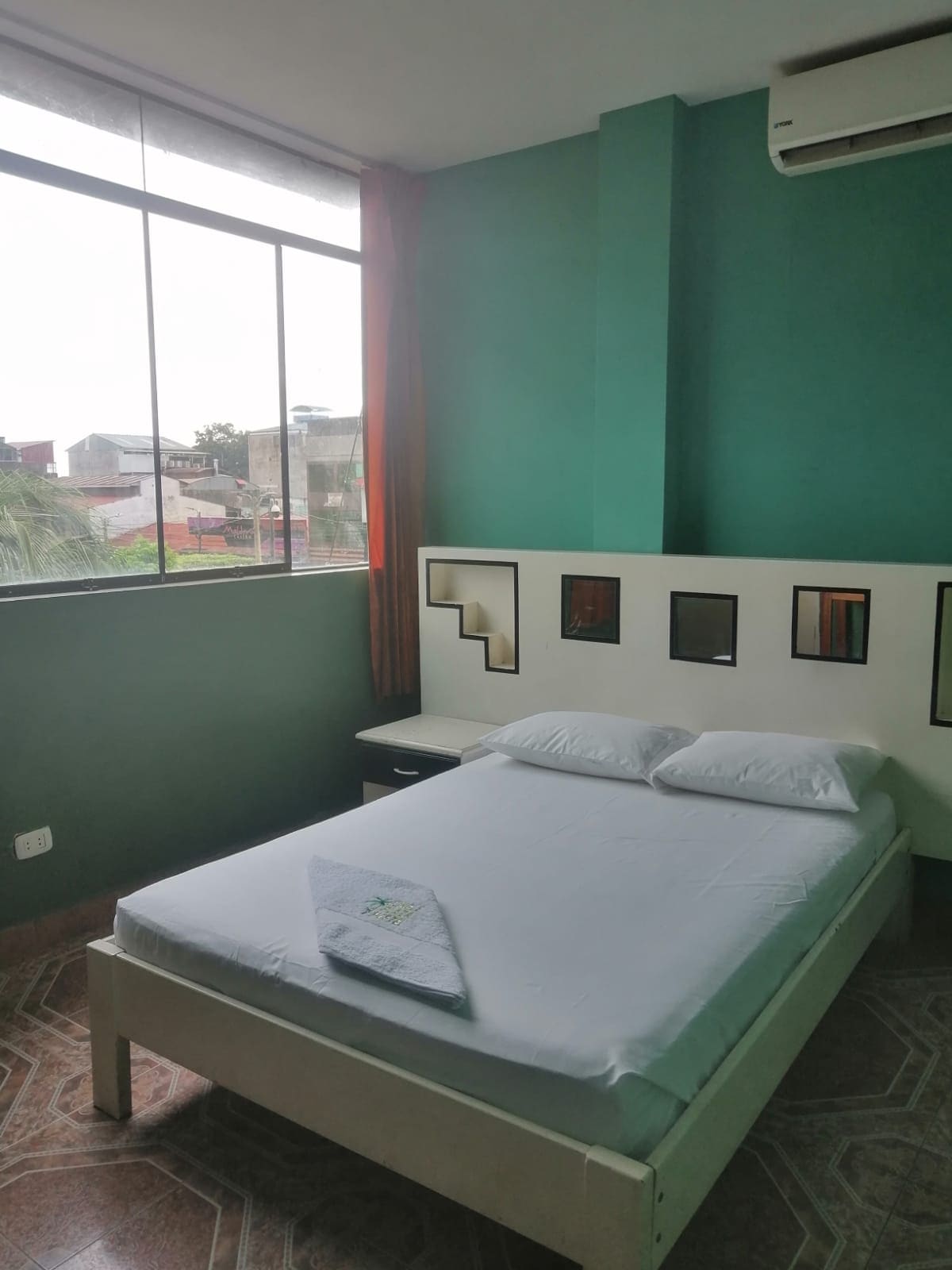 A well-lit sleeping area features a spacious bed with white bedding. A modern headboard adorned with decorative mirrors complements the green walls. Large windows allow natural light to fill the room, while a small bedside table is positioned next to the bed.