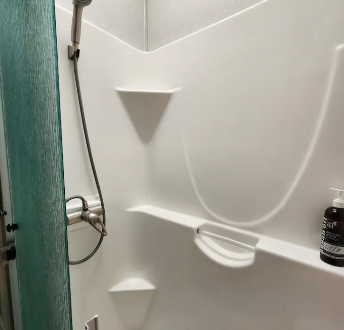 A modern shower area features smooth, white walls with built-in shelves for convenience. A handheld showerhead is attached to the wall, while a bottle of body wash is placed on one of the shelves. Light reflections create soft shadows on the surfaces.