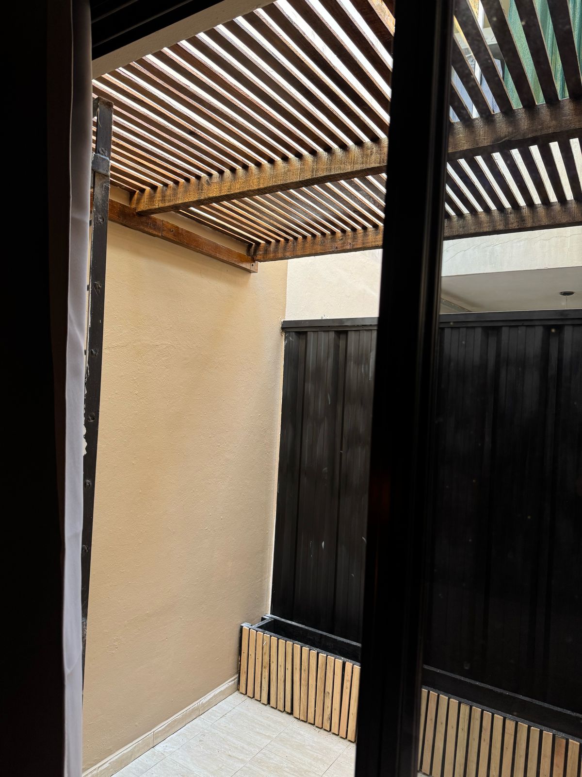 An outdoor area is shown, featuring a covered section with horizontal wooden slats providing partial shade. The walls are painted a soft beige, and a dark-finished fence creates a private atmosphere. Tiled flooring is visible under the slatted roof.