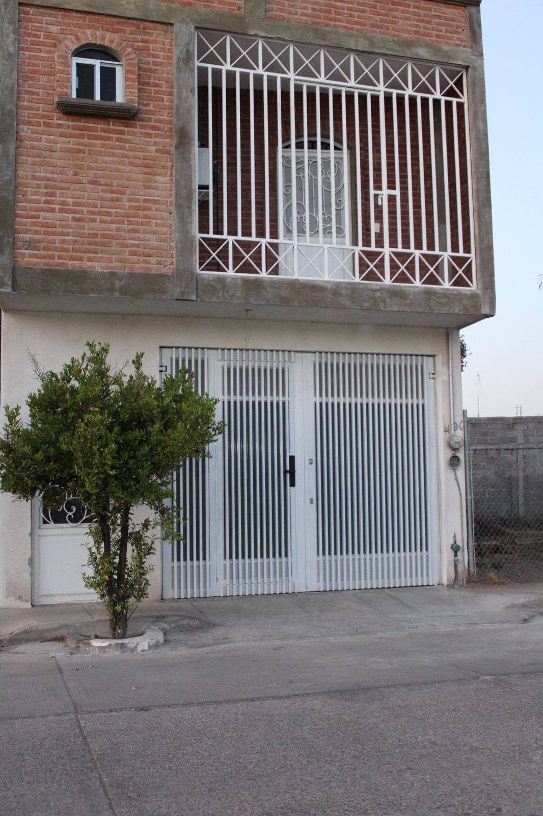 The exterior of a two-story building is shown, featuring a white gated entrance with vertical bars. A small tree is visible in front, and a door is framed by decorative metalwork. The building's brick upper level contrasts with the smooth white lower section.