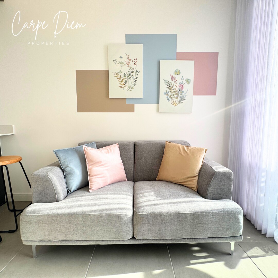A light grey sofa is positioned against a soft, neutral wall featuring decorative botanical prints. Two accent pillows in pastel colors are placed on the cushions, adding subtle pops of color. Natural light filters through sheer curtains, illuminating the cozy seating area.