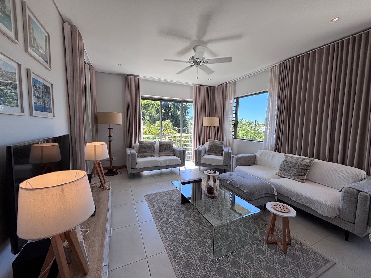 A spacious living area features large windows allowing for natural light. Two comfortable sofas are positioned around a glass coffee table, complemented by modern lamps. Soft grey curtains frame the windows, enhancing the room's tranquility.