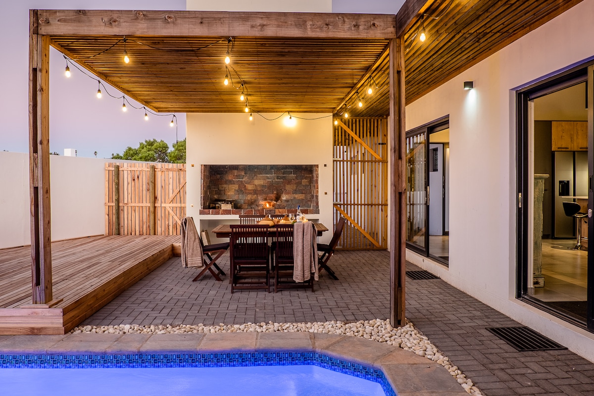 An outdoor dining space is featured under warm string lights, with a wooden table surrounded by matching chairs. A stone fireplace provides a central focal point, while a swimming pool is visible to one side, enhancing the setting for gatherings.
