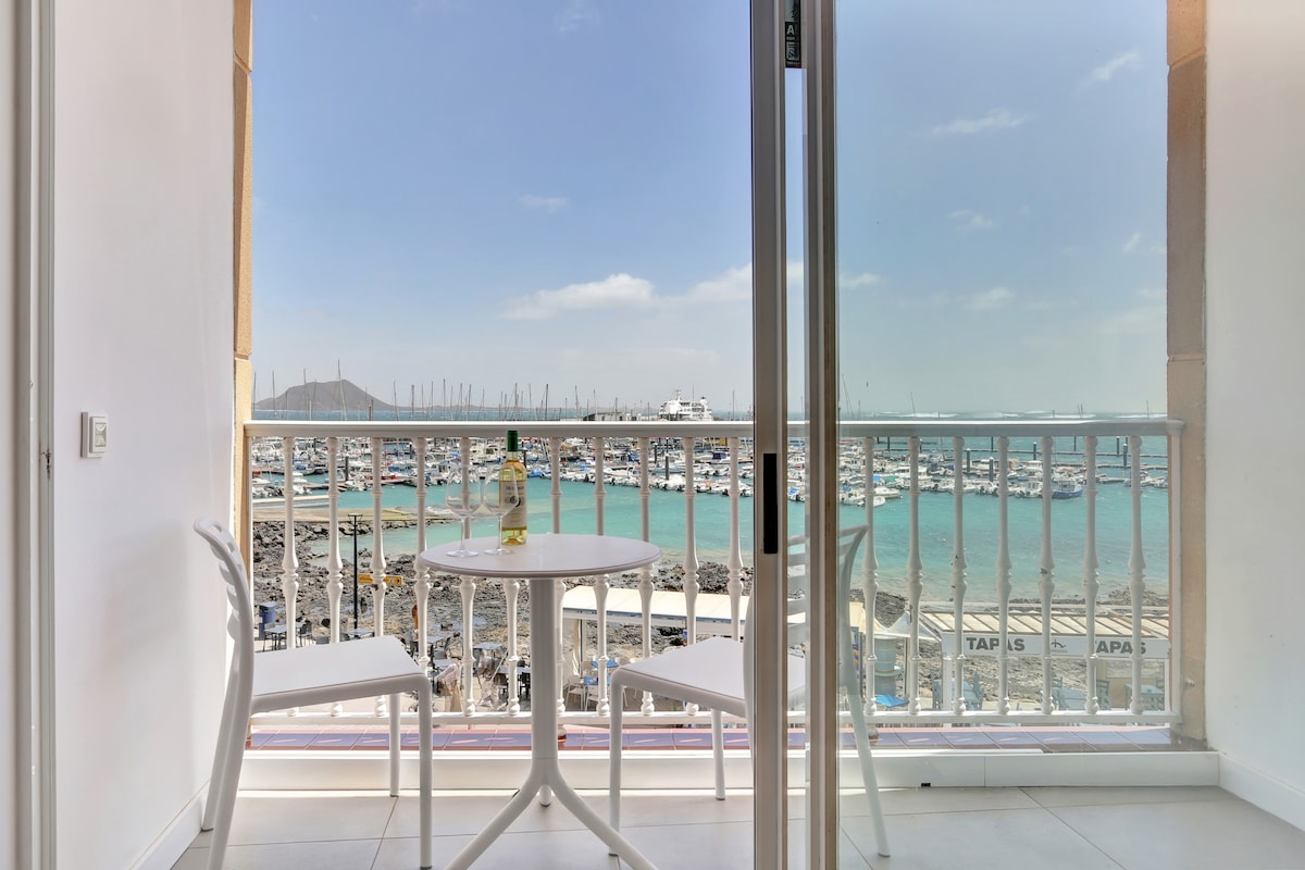 A private terrace is shown with a small round table and two white chairs, overlooking the marina. The view presents a vibrant marina filled with boats and distant island scenery, with blue waters reflecting the sky.