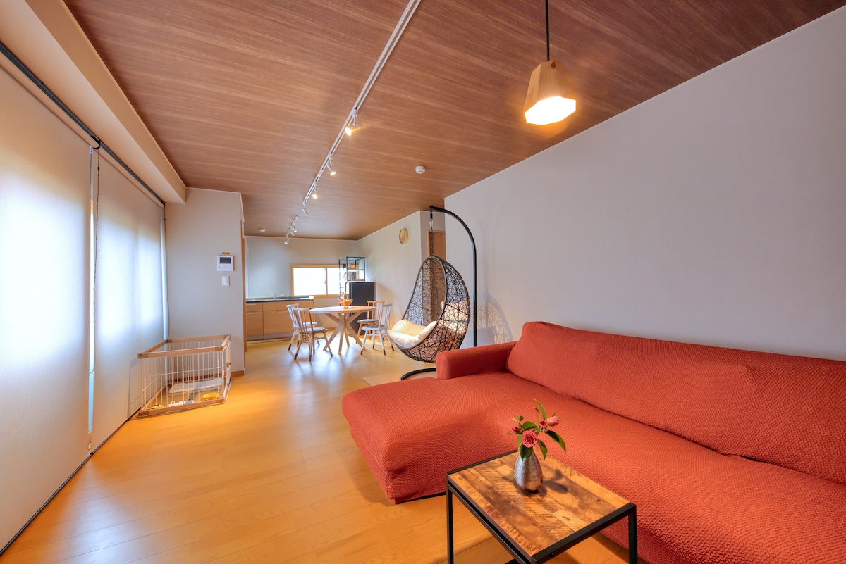 A spacious living area features a comfortable orange sofa and a hanging chair, creating a relaxed seating arrangement. A small table sits on a wooden floor, while a window allows natural light to brighten the space. A pet enclosure is also visible in the corner.