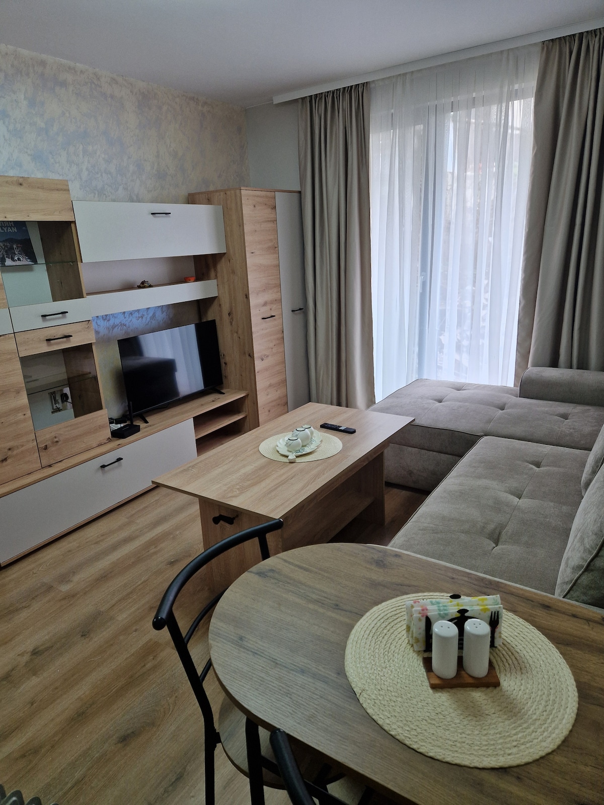 A cozy living area features a sectional sofa in a neutral tone and a wooden coffee table. Light-colored walls and curtains allow natural light to fill the space. A compact TV stand is positioned opposite the seating, adding functionality to the room.