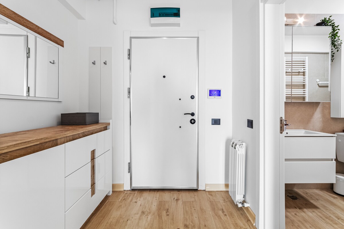A bright entrance area is shown, featuring a modern white door with a sleek design. A wooden console table with storage drawers is positioned against the wall. Mirrors and a small shelf are mounted nearby, complementing the contemporary decor and light wood flooring.