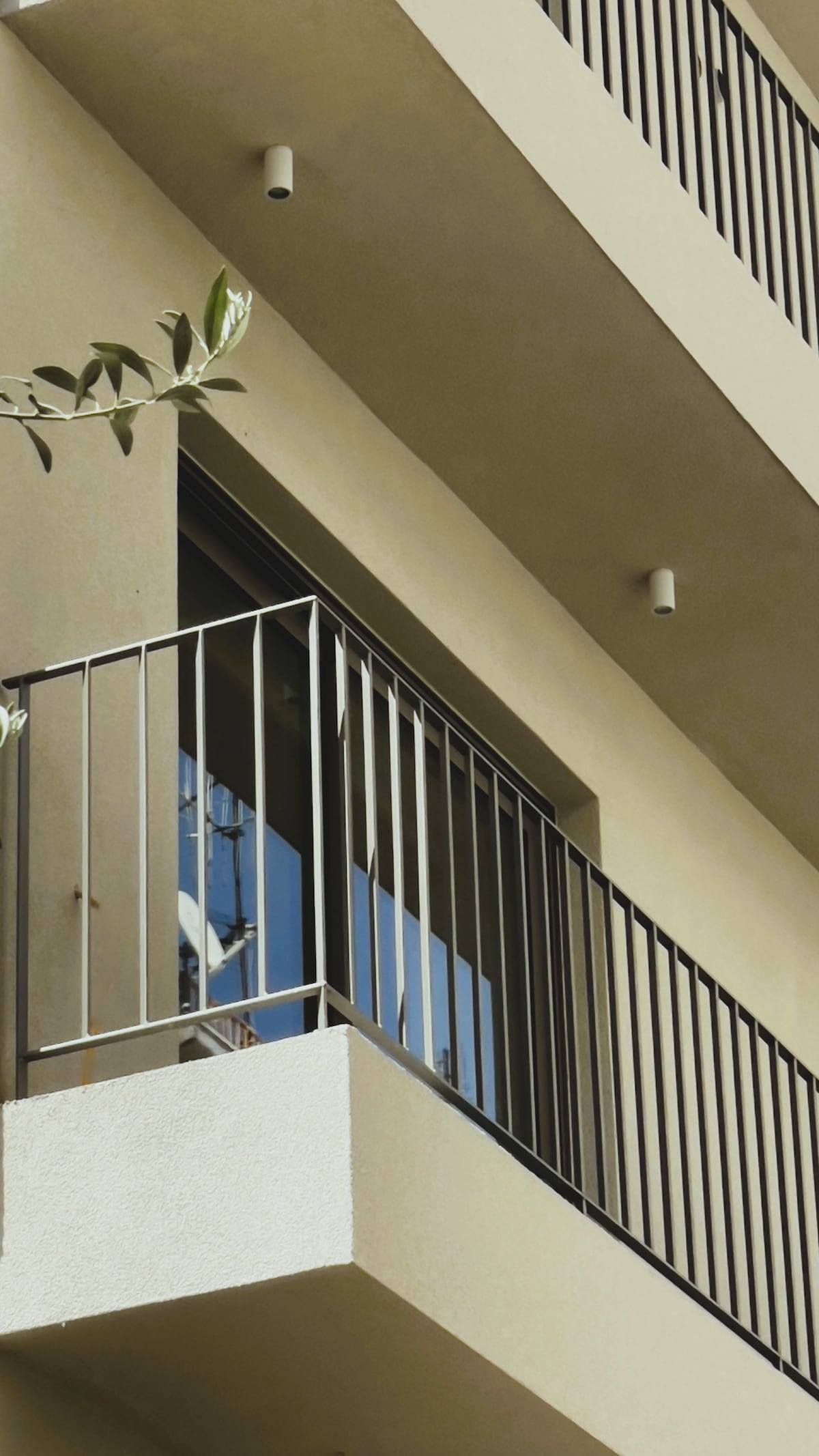 The image depicts a modern building exterior with a clean, minimalist aesthetic. A balcony with a sleek metal railing extends from the second floor, featuring glass doors that reflect nearby structures. Soft beige walls complement the design, providing a contemporary feel.