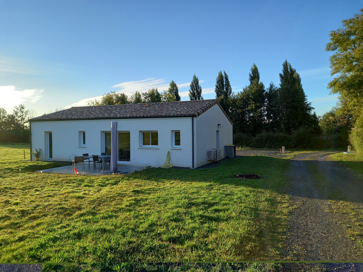 The exterior of a modern, single-story house is depicted, surrounded by green grass and trees. A patio area is shown with outdoor furniture. A gravel path leads to the entrance, reflecting the tranquil rural setting.