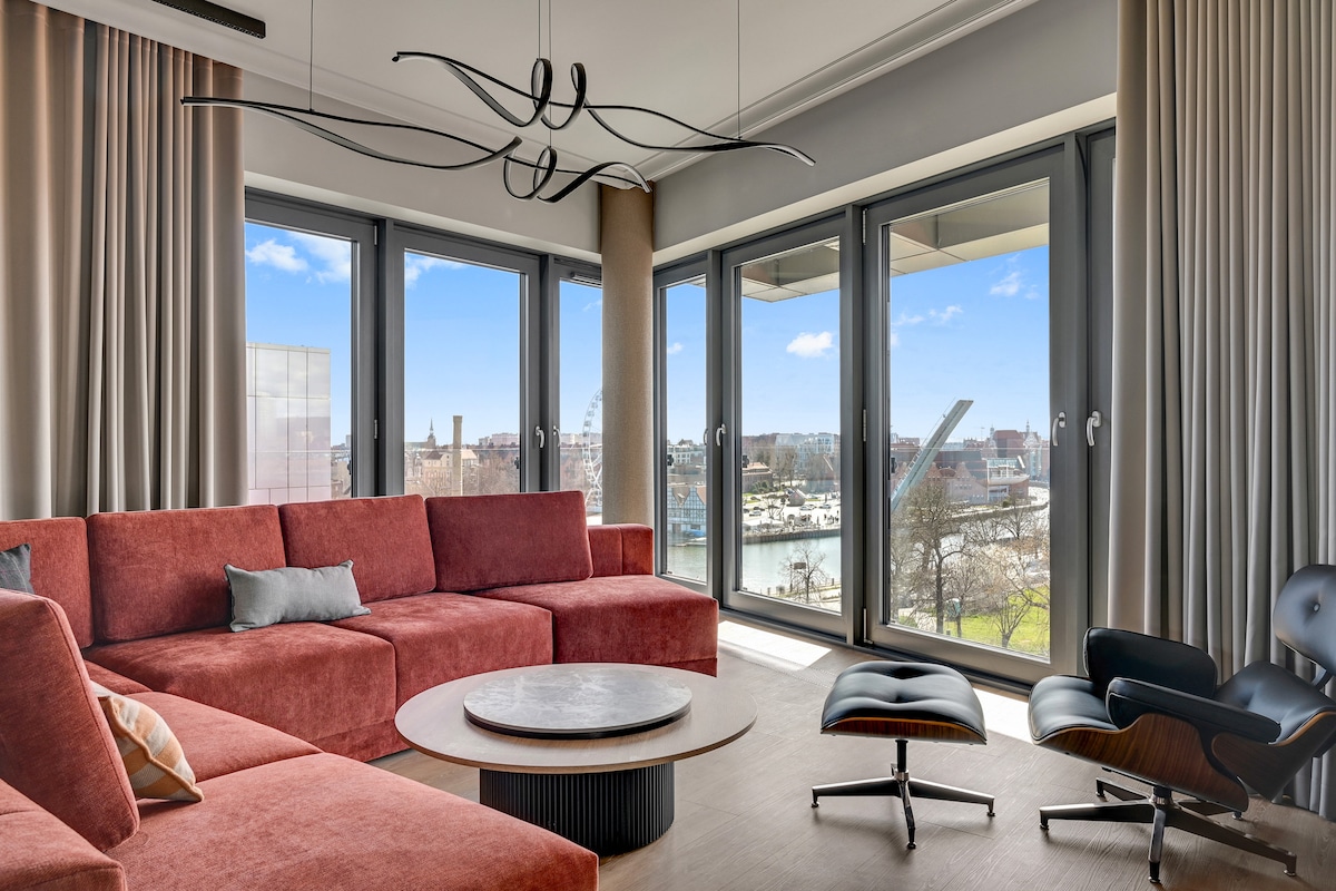 A spacious living area features a large, sectional sofa in warm tones, complemented by a round coffee table. Floor-to-ceiling windows provide abundant natural light and stunning views of the cityscape, including historic buildings and waterways. Modern light fixtures enhance the sleek design.