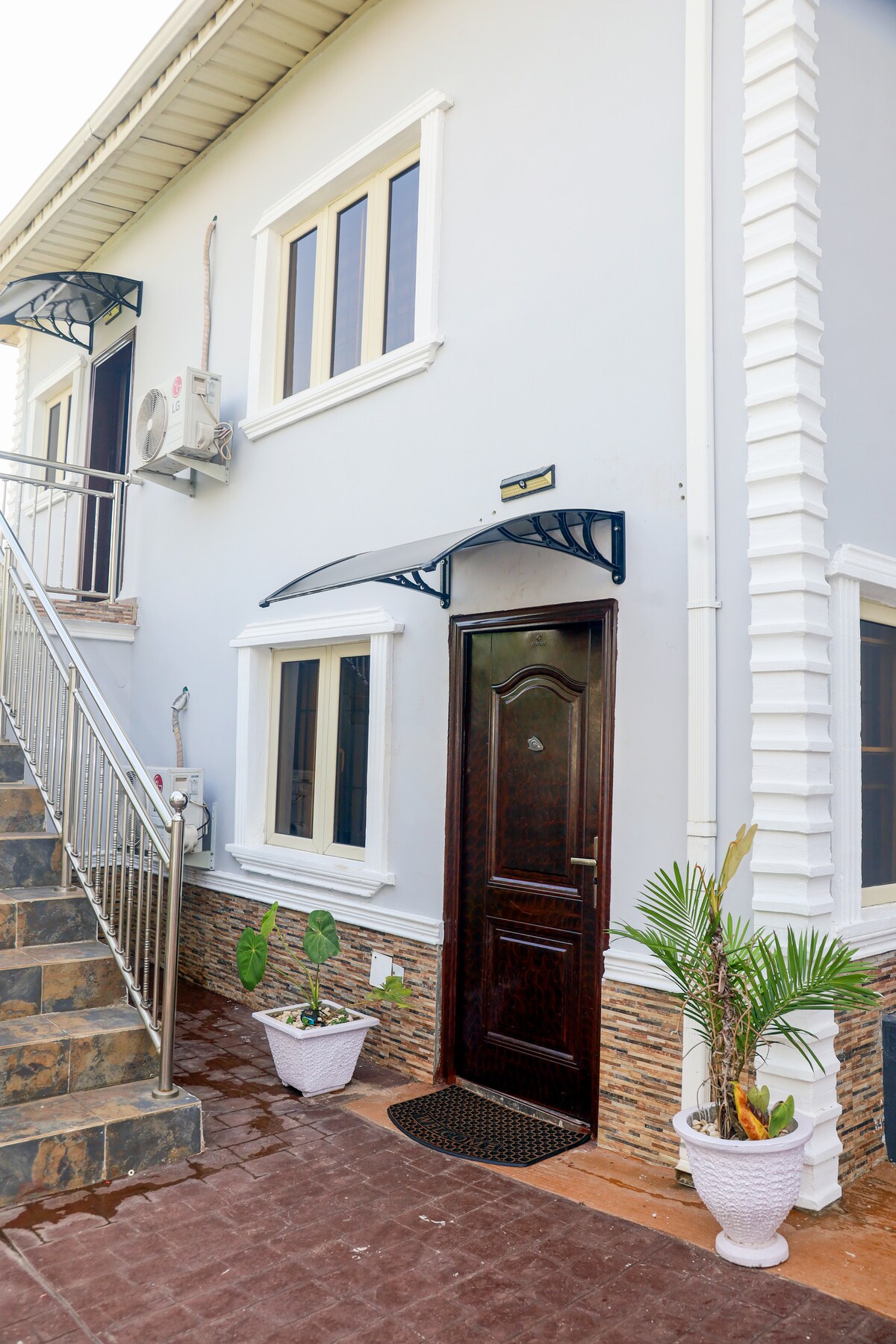 The exterior of the building features a well-maintained entrance framed by decorative elements. Two large windows and a door are seen, with plants placed in white pots on either side. A staircase leads to the upper level, enhancing accessibility.