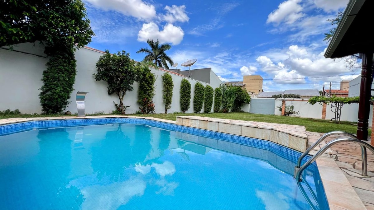 A tranquil swimming pool is surrounded by greenery, with clear blue water reflecting the sky. The pool area includes a small seating space and a stone edge, providing a relaxed outdoor setting. Lush trees and shrubs frame the area, contributing to a peaceful ambiance.