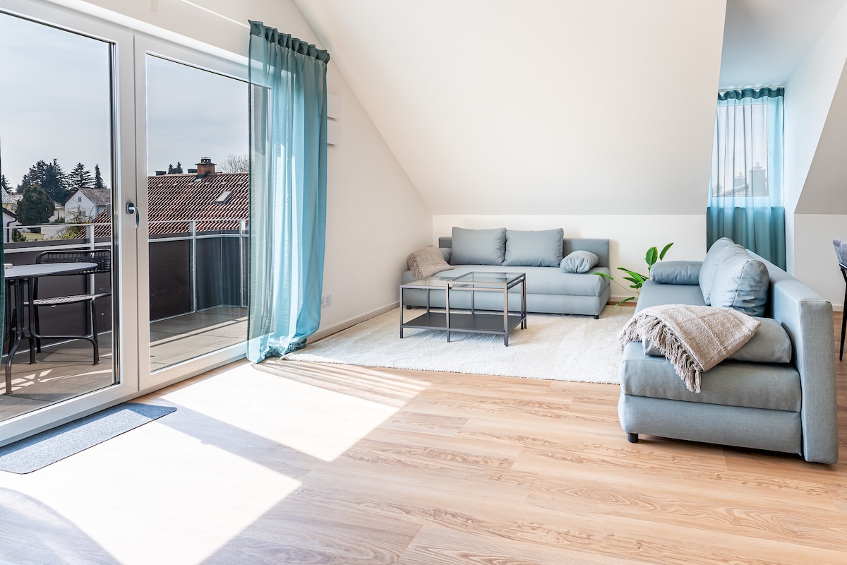 A bright and airy living area features large glass doors that open to a balcony, allowing natural light to fill the space. Two light blue sofas are arranged with a coffee table in between, complemented by light-colored wooden flooring.
