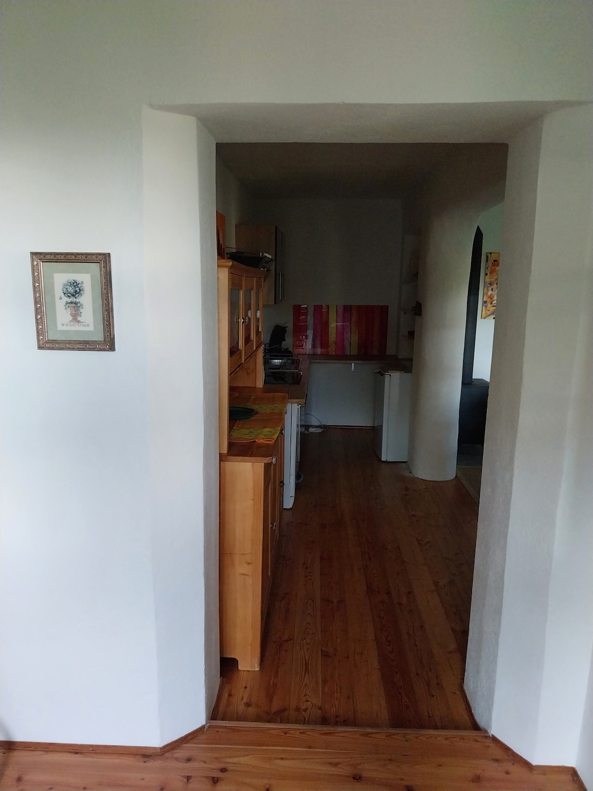 A corridor leads to a kitchen area with wooden cabinetry and decorative wall art. The warm wooden flooring enhances the space's inviting quality. At the end of the corridor, a glimpse of another room is visible, suggesting an open and connected layout.