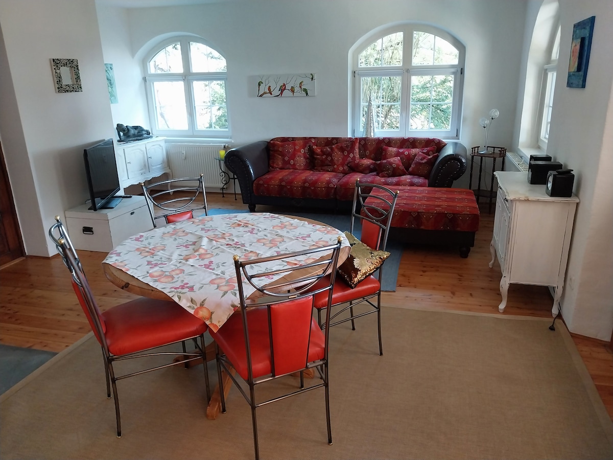 A spacious living area features a patterned red couch and a dining table set with four silver metal chairs. Large windows allow natural light to fill the room, offering views of the greenery outside. A small television is positioned near the seating area.