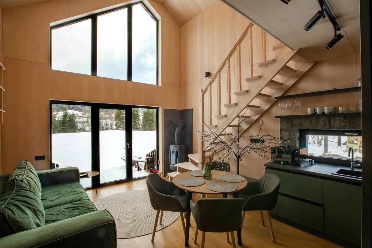 The open living area is highlighted by large windows, allowing natural light to fill the space. A cozy green sofa is paired with a circular dining table and four chairs. A staircase leads to the upper level, while a fireplace is visible in the corner.