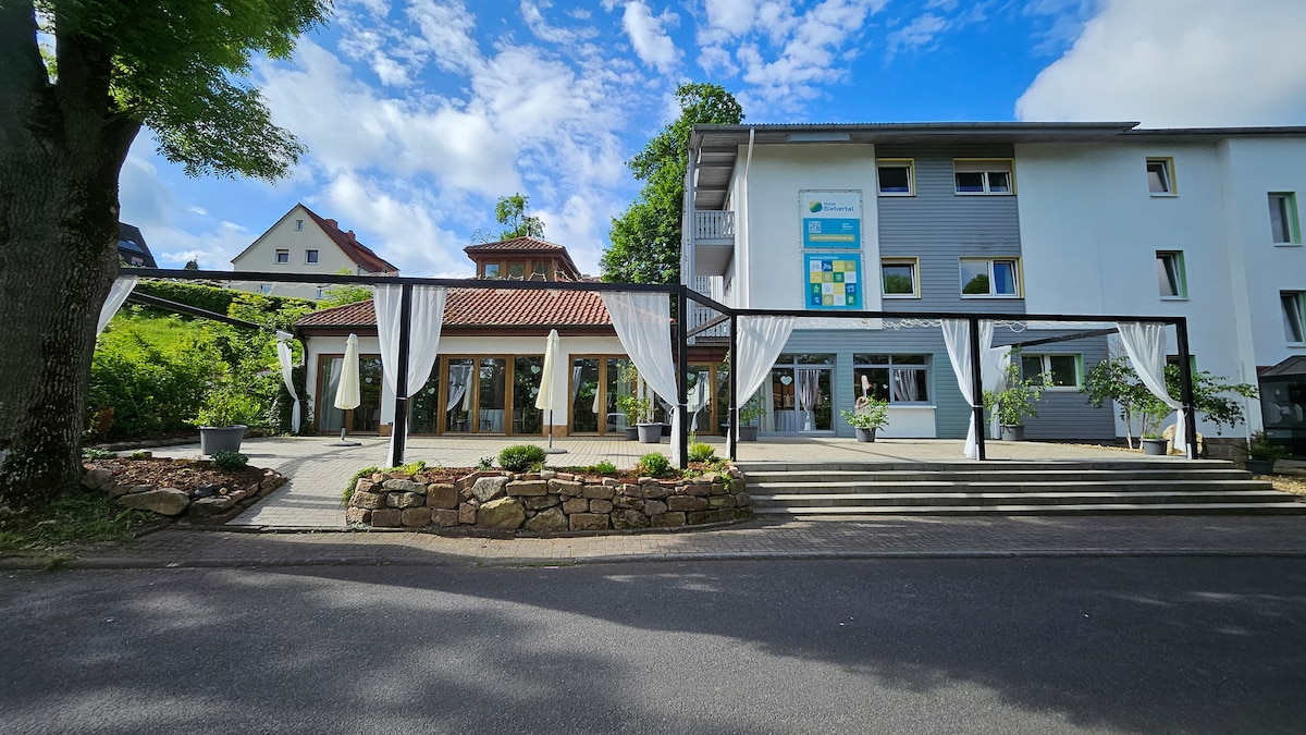 The exterior of a hotel is shown with a welcoming entrance framed by decorative awnings. A patio area features several tables with umbrellas, surrounded by neatly arranged greenery and stone landscaping. The building's architecture includes a combination of modern and traditional elements under a partly cloudy sky.