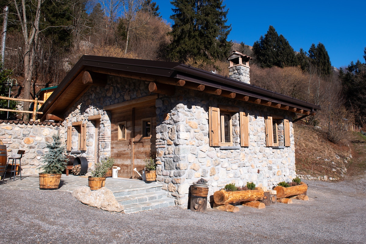 A charming stone structure surrounded by nature, featuring wooden shutters and a cozy chimney. The entrance includes planters with greenery and decorative barrels, while gravel pathways lead to the inviting facade, all set against a backdrop of trees and open land.