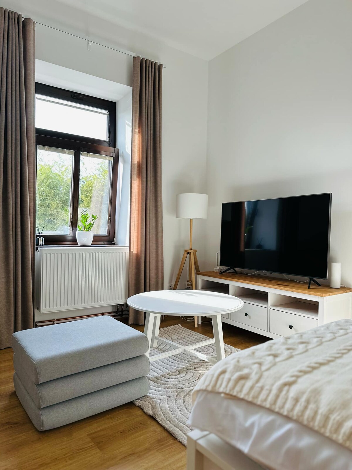 A well-lit corner of a living area is depicted, featuring a large television mounted on a light wooden stand. A round white table is positioned in front of a cozy gray ottoman. Natural light filters through a window dressed with long, beige curtains.
