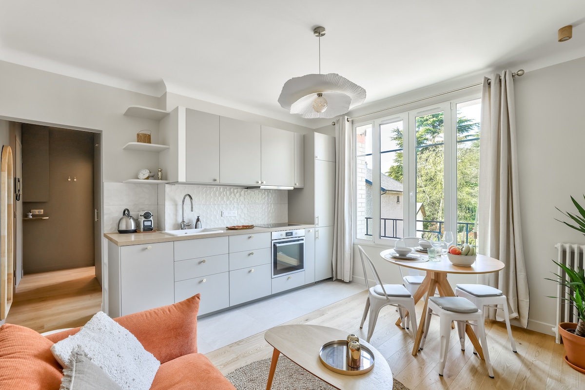 A modern kitchen and dining area are showcased, featuring light cabinetry and an integrated oven. Natural light streams through large windows, illuminating a round dining table surrounded by wooden chairs. A cozy seating arrangement is visible, complemented by soft textiles and greenery.