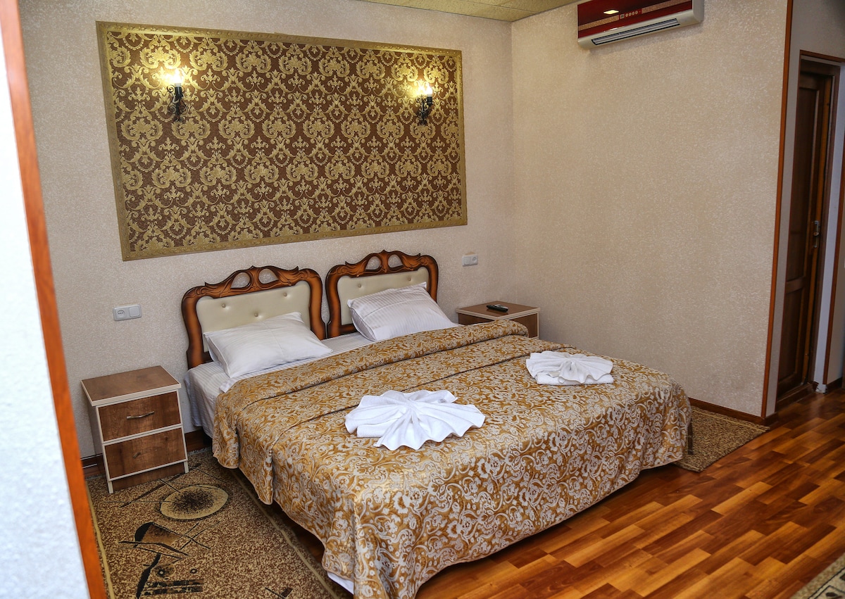 A sleeping area features a spacious bed covered with a decorative gold-patterned blanket. Two bedside tables are placed on either side, and a subtle wall design adds visual interest. Soft lighting is provided by wall sconces, and wooden flooring completes the warm ambiance.