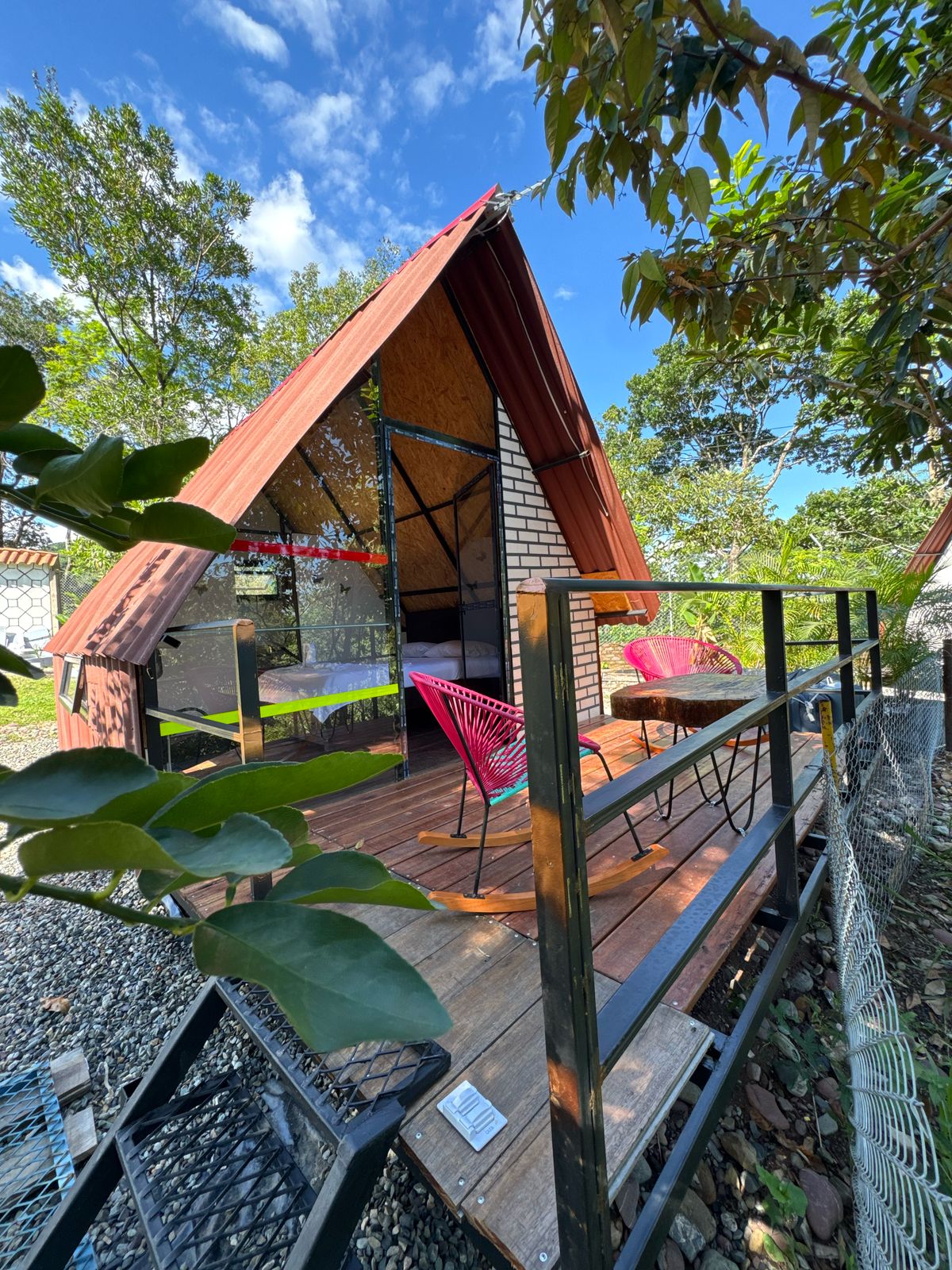A triangular cabin features a wooden deck with two pink chairs and a small table set against a backdrop of lush greenery. Natural light is captured through large glass windows, and the cabin's metal roof complements the wooden elements.