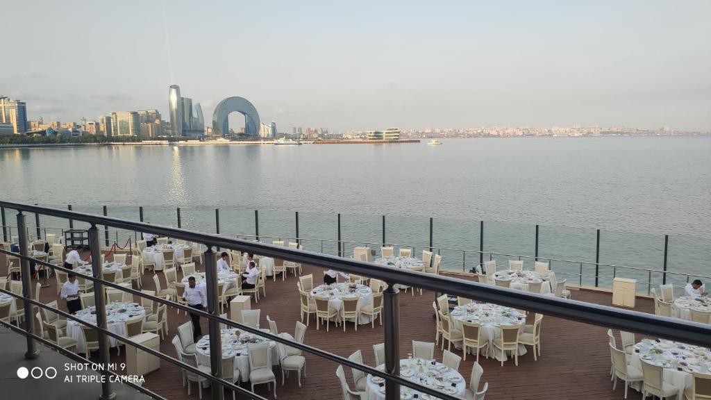 A spacious outdoor dining area is seen with elegantly set tables featuring white tablecloths and matching chairs. The view overlooks the Caspian Sea, with a distant skyline including modern architecture and a distinctive arch-shaped structure.