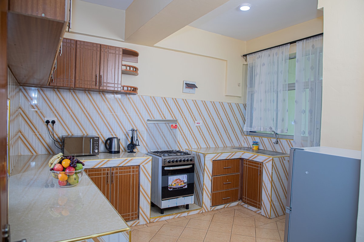 A functional kitchen space features patterned cabinetry and a light-colored countertop. Modern appliances, including a stove and microwave, are visible, along with a refrigerator. A bowl of fresh fruit is placed on the counter, and natural light filters through a partially draped window.