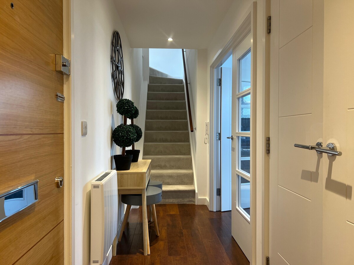 A well-lit entryway showcases a wooden door with a modern mailbox. A small table is placed against the wall, accompanied by two decorative potted plants. Staircase leads to the upper level, with plush carpeting and natural light filtering through glass doors.