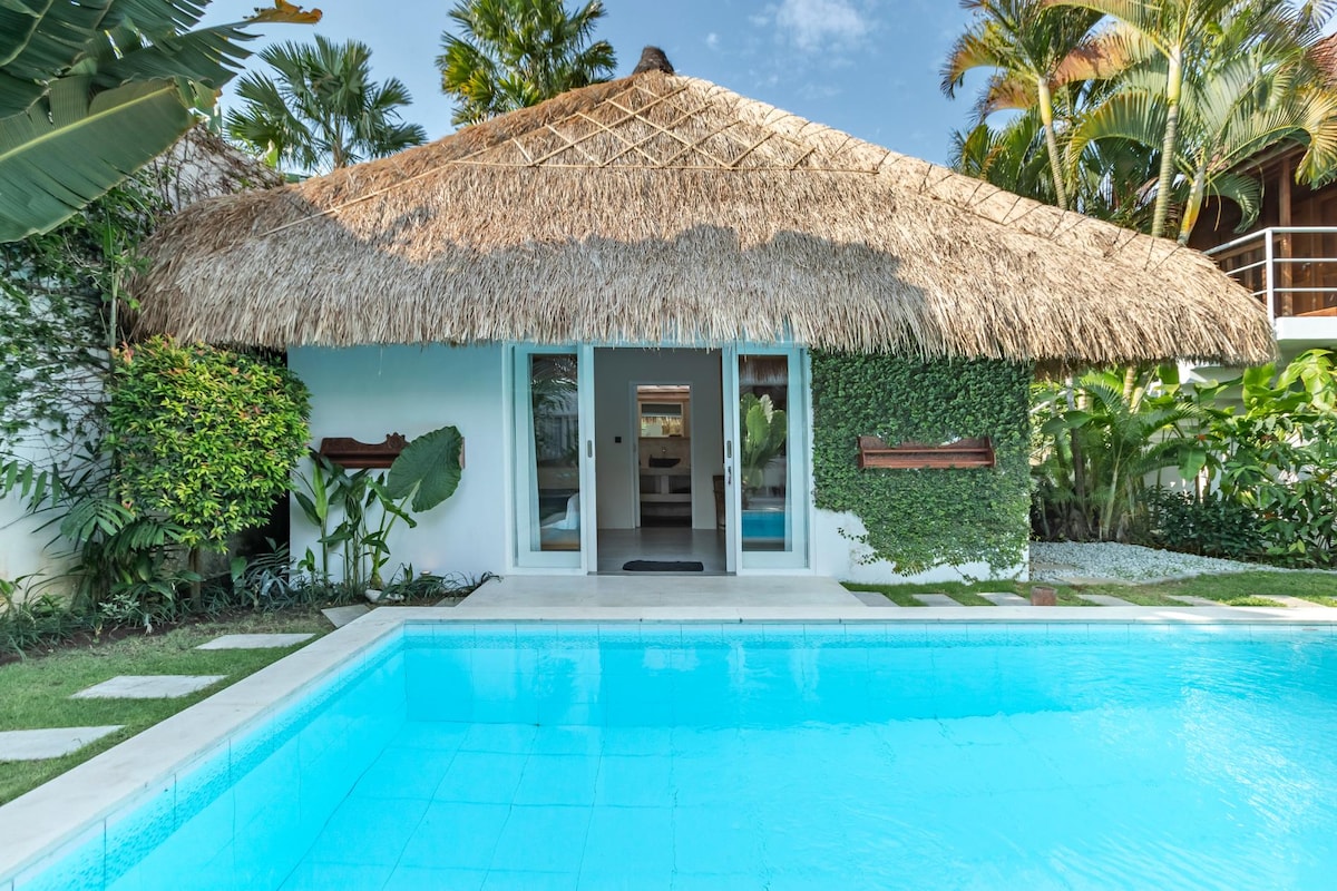 A Joglo-style villa is set amidst lush gardens and palm trees, showcasing a thatched-roof exterior. Large glass doors open toward the turquoise pool, creating a seamless connection between indoor and outdoor spaces. Natural greenery adorns the walls, enhancing the tranquil ambiance.