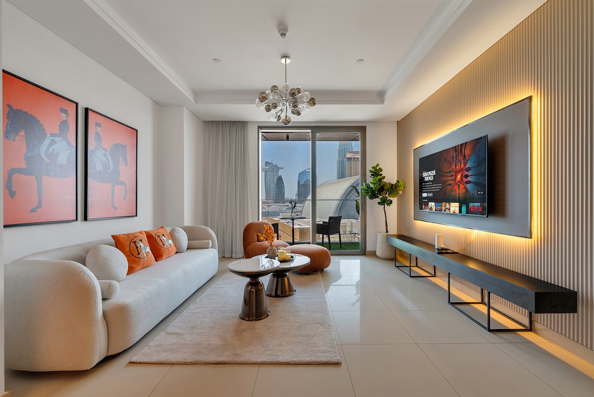 A living area showcases a cozy, curved sofa paired with plush orange cushions. A sleek coffee table and two metallic side tables occupy the space. A large smart TV is mounted on a stylish wall, and floor-to-ceiling windows provide a view of the city skyline.