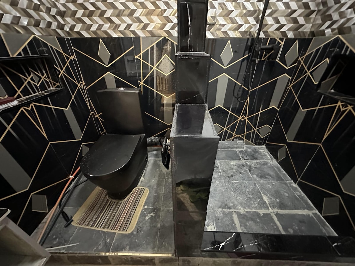 A modern bathroom is displayed, featuring dark tiles with geometric patterns. A black toilet is positioned adjacent to a stylish shower area with a sleek black partition. A small mat adds comfort to the space, while shelves provide storage for toiletries.
