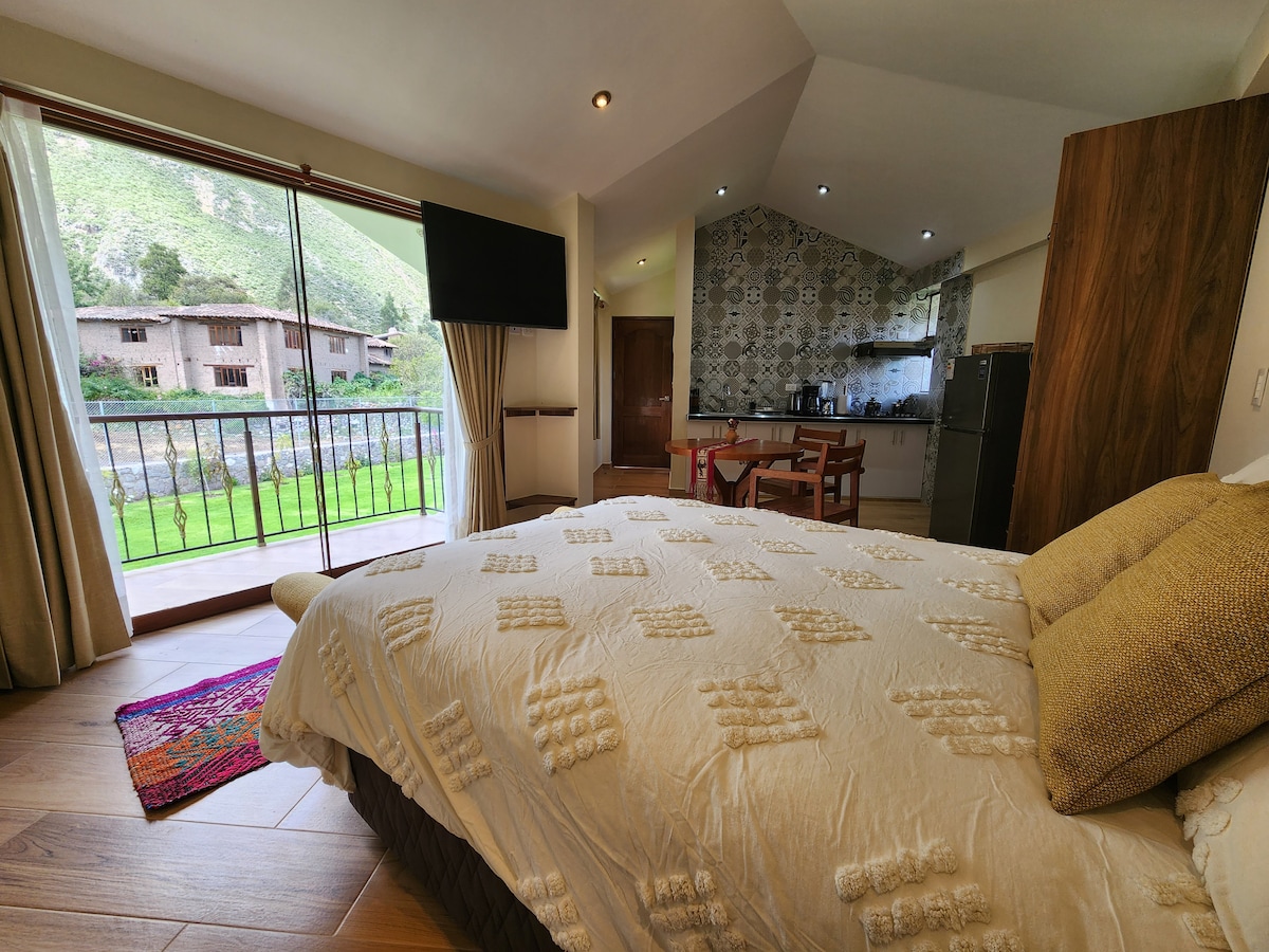 A cozy sleeping area features a spacious bed adorned with a textured duvet. Large glass doors open onto a private balcony, providing views of the landscaped garden and nearby mountains. A dining area with wood furniture and a kitchenette is visible in the background.