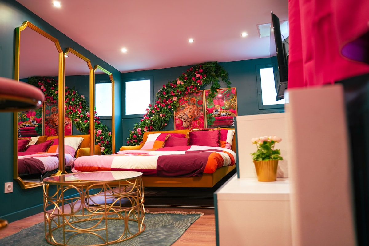 A modern studio bedroom features a spacious bed adorned with colorful pillows and bedding. A decorative wall of flowers frames the headboard, while a circular coffee table adds an elegant touch. Two large mirrors reflect the vibrant decor, and natural light enters through small windows.