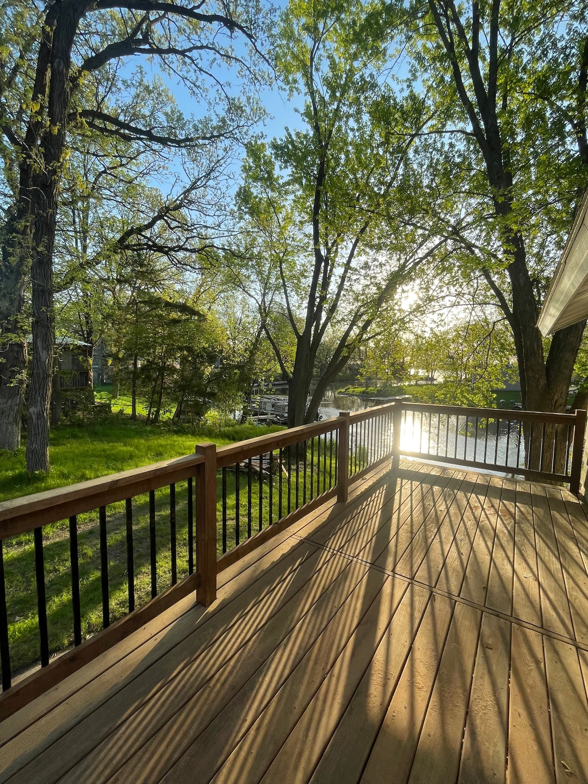 A spacious wooden deck features a railing along its perimeter, with sunlight filtering through the surrounding trees. The deck overlooks a tranquil body of water, creating a serene outdoor space perfect for relaxation.