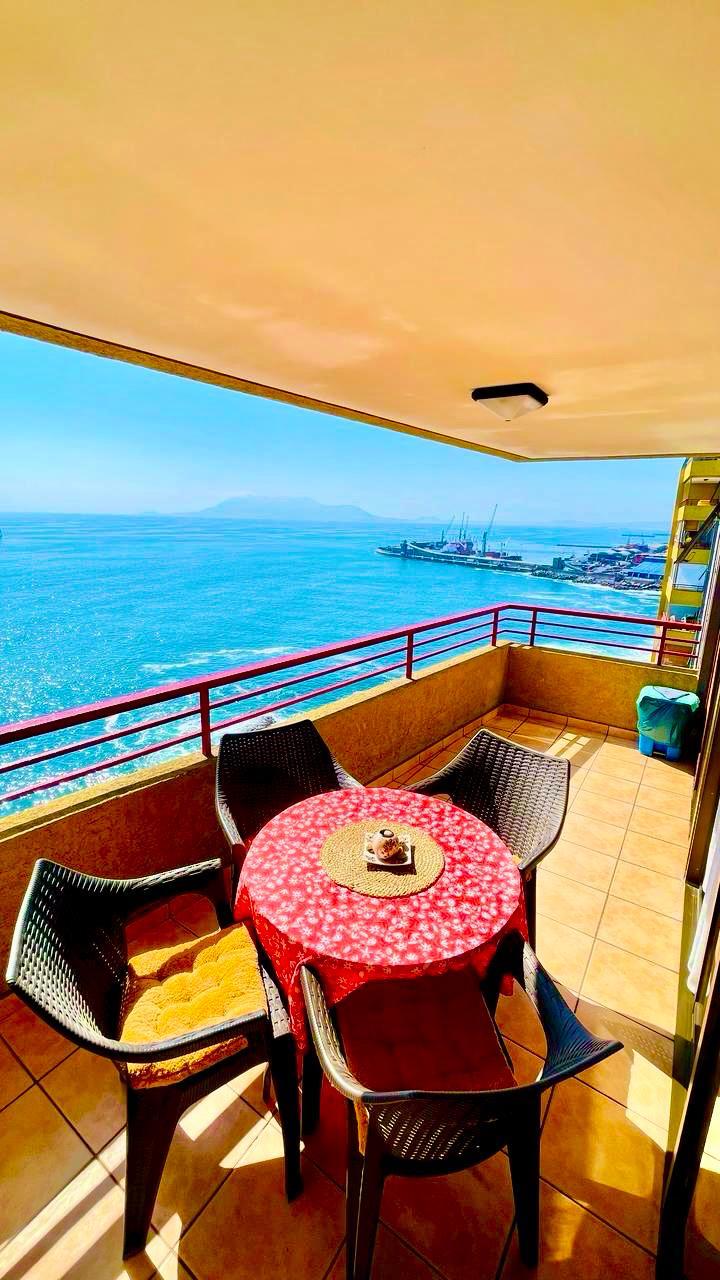 A terrace is shown overlooking the ocean, featuring a round table draped with a red tablecloth and surrounded by four black chairs. The expansive view reveals calm waters and distant boats, creating a relaxed outdoor space for enjoying the scenery.