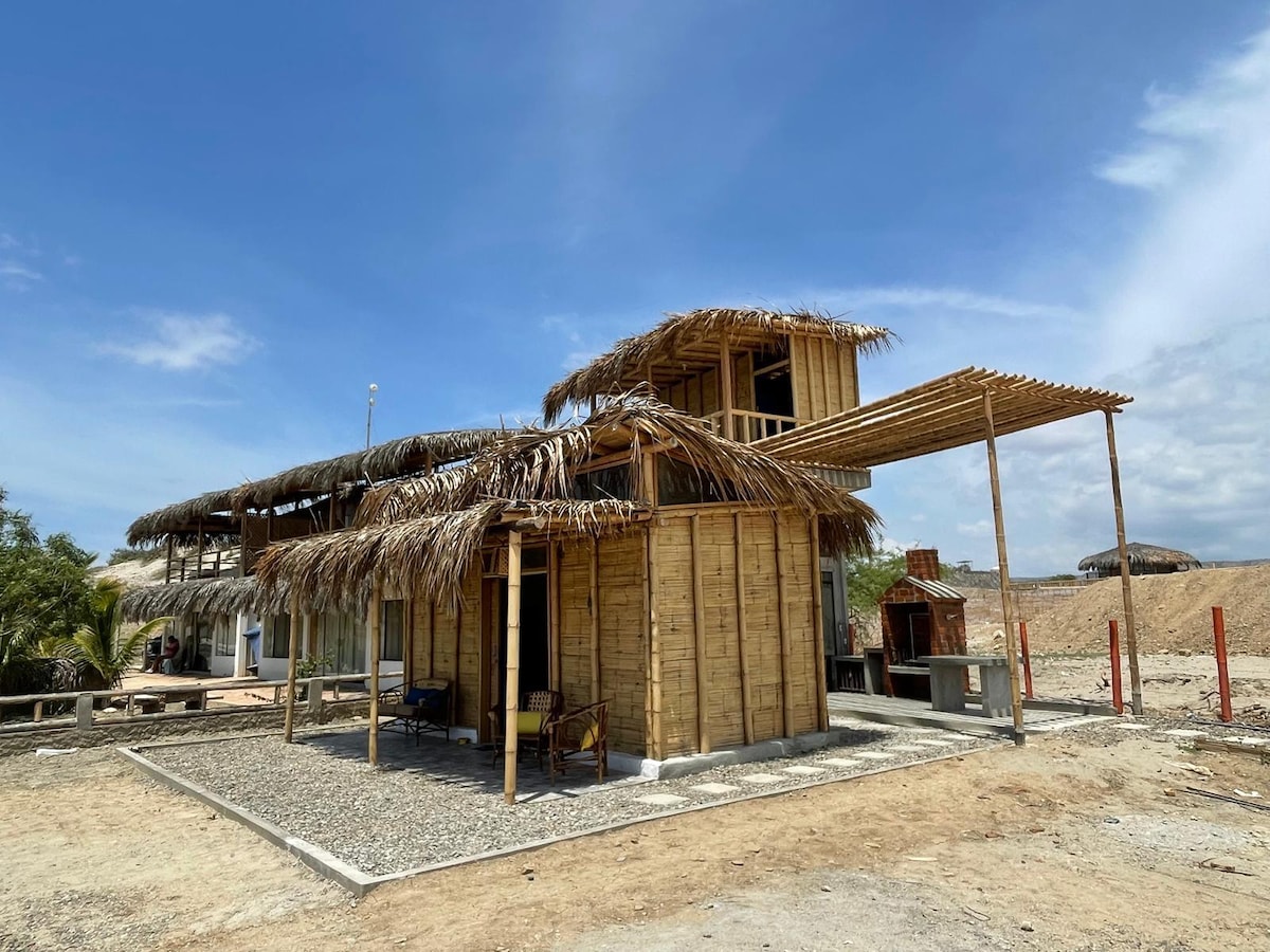 A spacious structure with a natural wood exterior and a thatched roof is set against a clear blue sky. The design features open spaces, wooden accents, and a combination of outdoor and shaded areas, integrating well with the surrounding landscape.