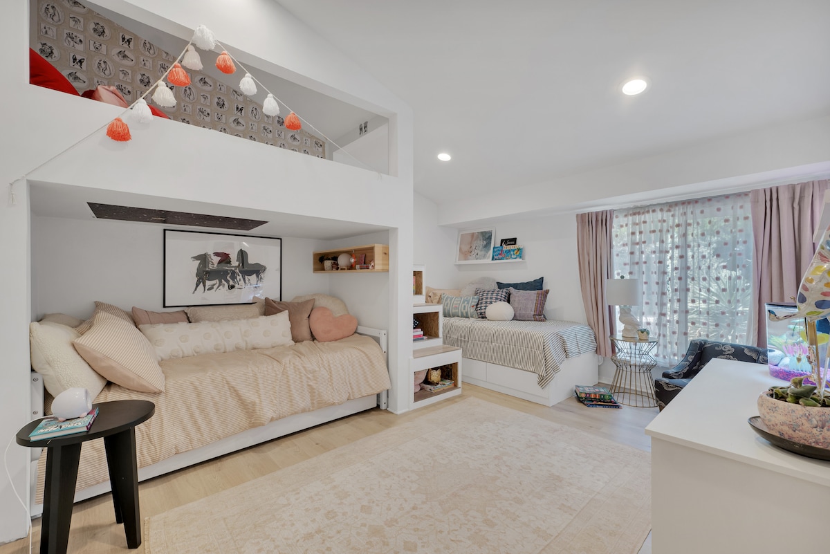 A bright and airy multi-purpose space features a lofted sleeping area above a cozy daybed adorned with soft pillows. A second bed sits nearby, complemented by natural light filtering through patterned curtains. Shelving displays decorative items, providing a casual yet inviting atmosphere.