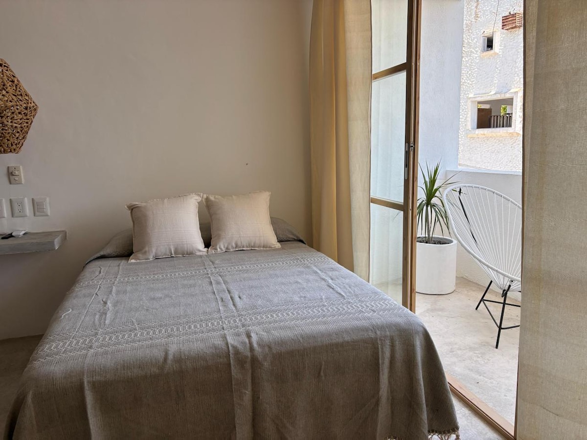 A modest bedroom features a spacious bed dressed in a gray blanket. Soft light enters from a sliding glass door that opens to a small balcony, where a potted plant and a white chair are positioned. Light-colored curtains frame the doorway.