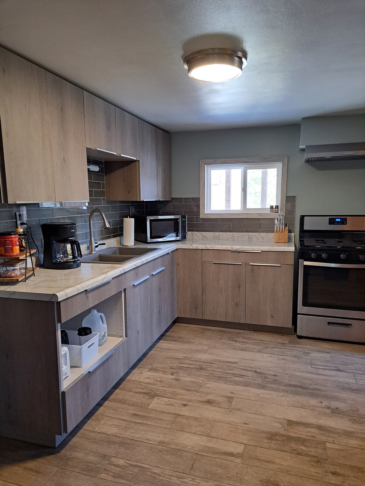 The kitchen features modern cabinetry in light wood tones, complemented by a marble countertop. A window offers natural light above the sink. Appliances include a stove, microwave, and coffee maker. The flooring is a warm, wood-like laminate, enhancing the spacious feel.