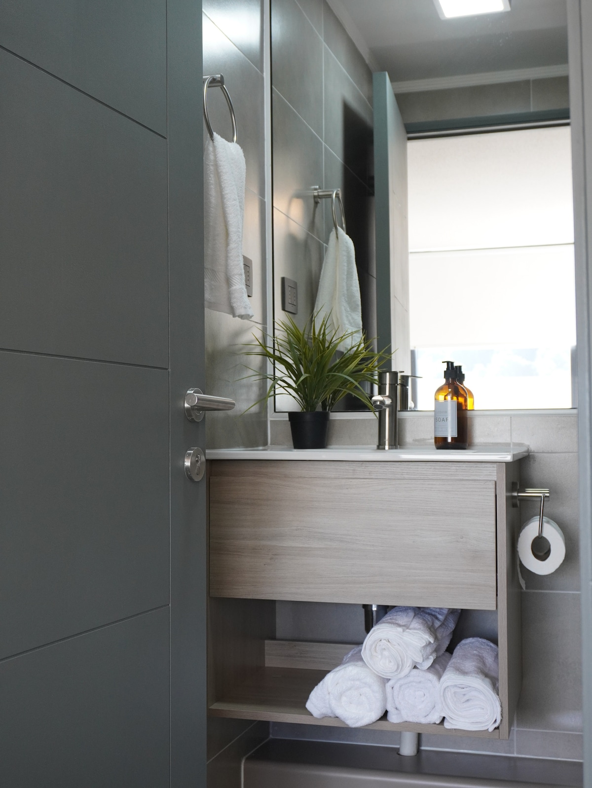 A modern bathroom is featured, showcasing a sleek vanity with a plant and toiletries. Fresh white towels are neatly arranged on a shelf below, while a mirror reflects the clean lines of the minimalist design. Soft light enhances the inviting atmosphere.