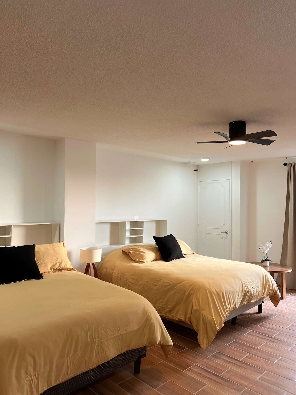 A spacious room is featured with two large beds dressed in golden-yellow linens. Each bed is accompanied by a bedside lamp and a dark cushion. A ceiling fan hangs above, and natural light enters through a nearby window, complemented by soft curtains.