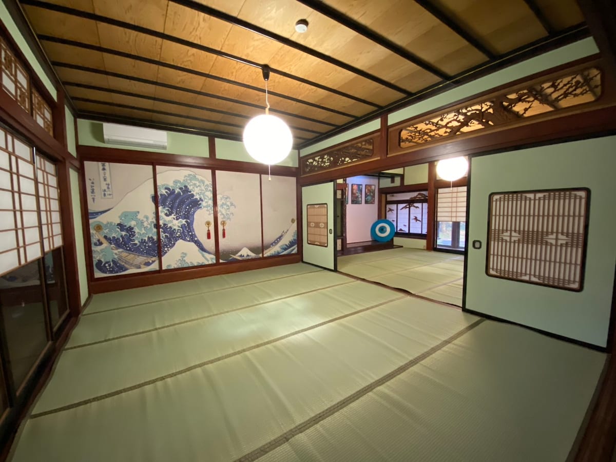 A traditional tatami room is featured, showcasing sliding doors and stylized wall art depicting waves. Natural wood beams highlight the ceiling, while soft lighting creates an inviting space. Multiple areas with tatami mats are available for seating or resting.