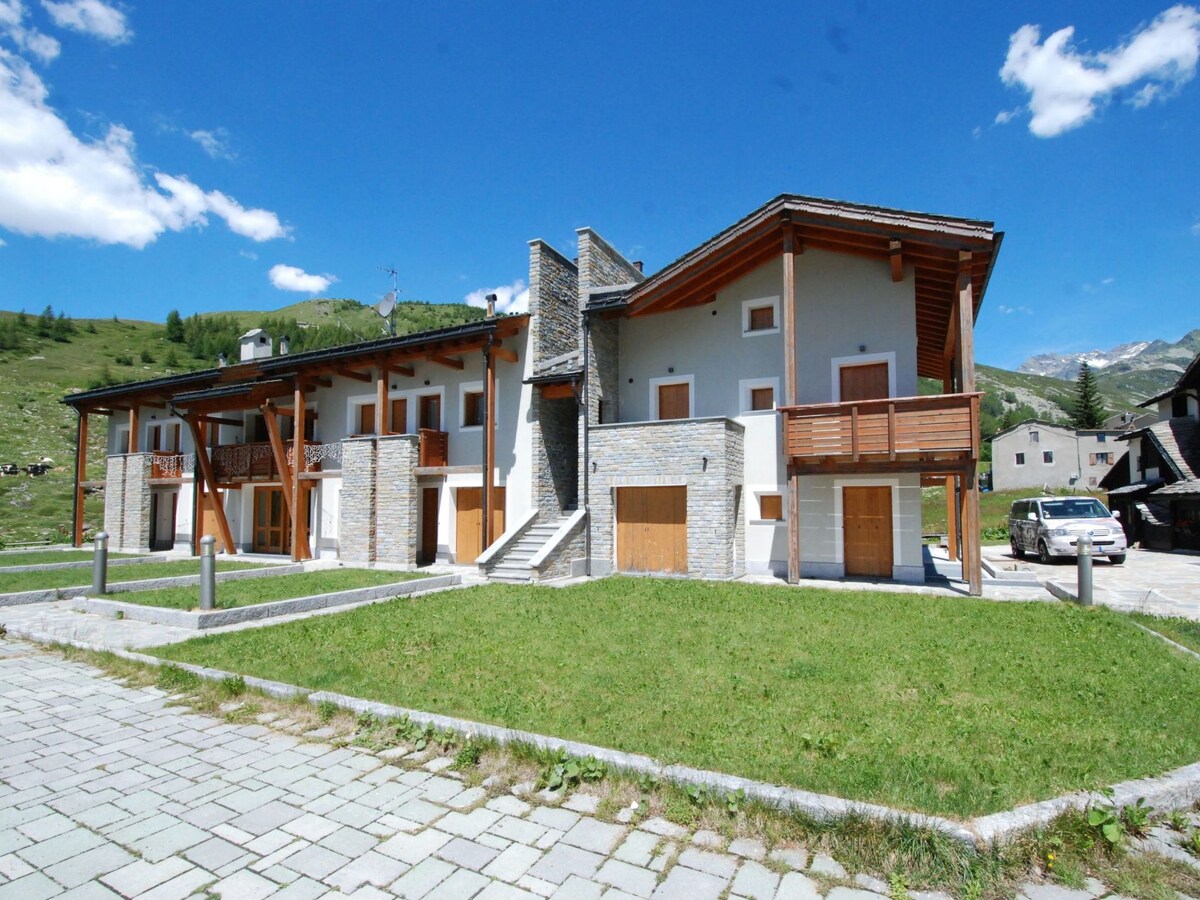 The exterior of the residence is depicted, featuring a two-story structure with a combination of stone and wooden elements. Large balconies and entrance doors are visible, surrounded by a grassy area. The scenic hillside in the background is complemented by a clear blue sky.