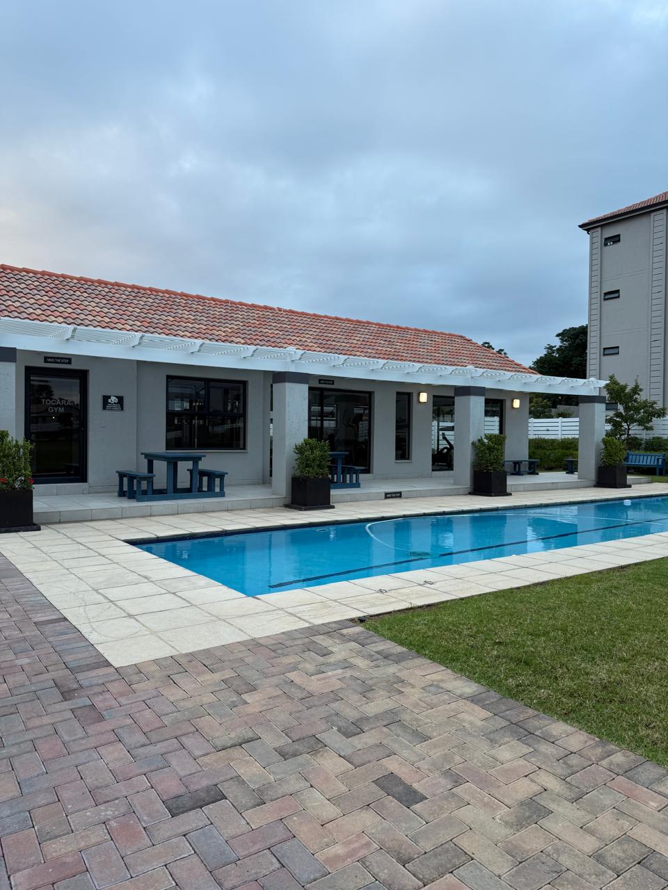 A modern exterior features a swimming pool set adjacent to a simple patio area. Potted plants flank the entrance, while a shaded area with tables and chairs invites outdoor relaxation. The building's clean lines and red-tiled roof add to the overall contemporary charm.