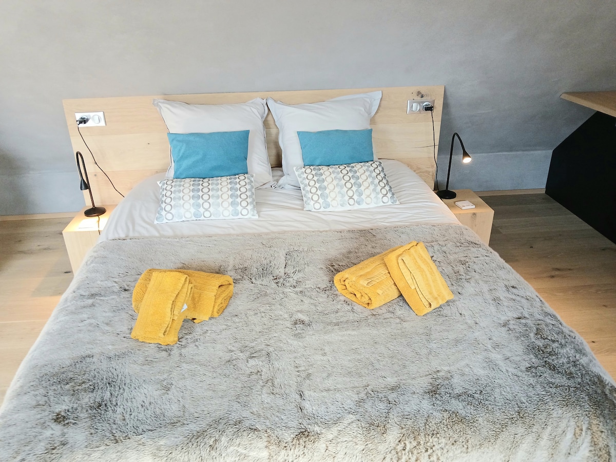 A spacious bed is adorned with white pillows and light blue accent cushions. A textured grey throw is draped across the bed, and two neatly rolled yellow towels are placed on top. Soft lighting is provided by wall-mounted lamps on either side.