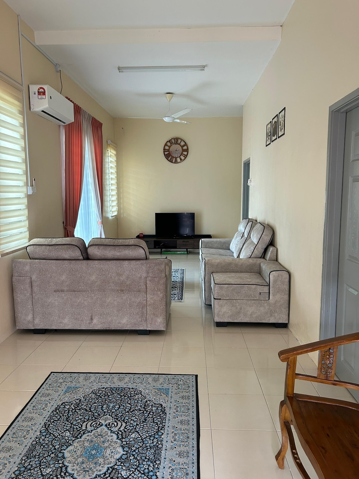 A living area is characterized by light-colored walls and tiled flooring. Two comfortable couches face a television set, with a wall clock above. Natural light enters through large windows, partially covered by sheer curtains. A decorative rug enhances the space, adding texture.